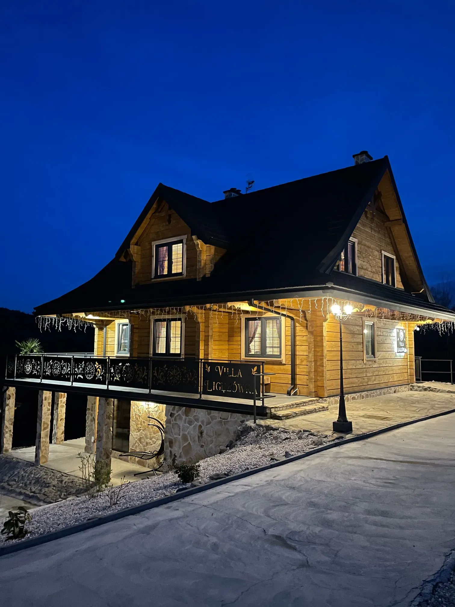 Drewniany dom z podświetlonym gankiem i kutą balustradą tarasu, wieczorem, z tablicą z nazwą obiektu