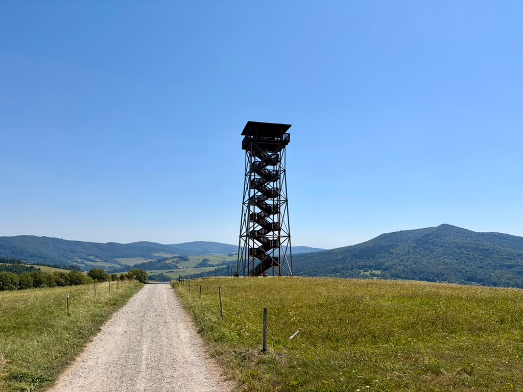 Droga szutrowa między łąkami prowadzi na grzbiet, tuż obok wieży: ciemnej stalowej kratownicy ze schodami biegnącymi zygzakiem na platformę pod dachem