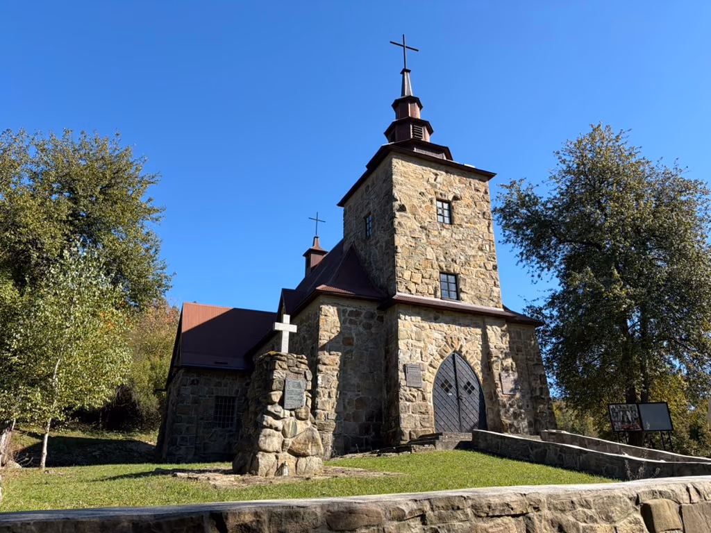 Kamienna fasada z kwadratową wieżą i krzyżem na iglicy, ostrołukowe drzwi, a przed wejściem kamienny słup z białym krzyżem; po bokach drzewa, nad nimi bezchmurne niebo