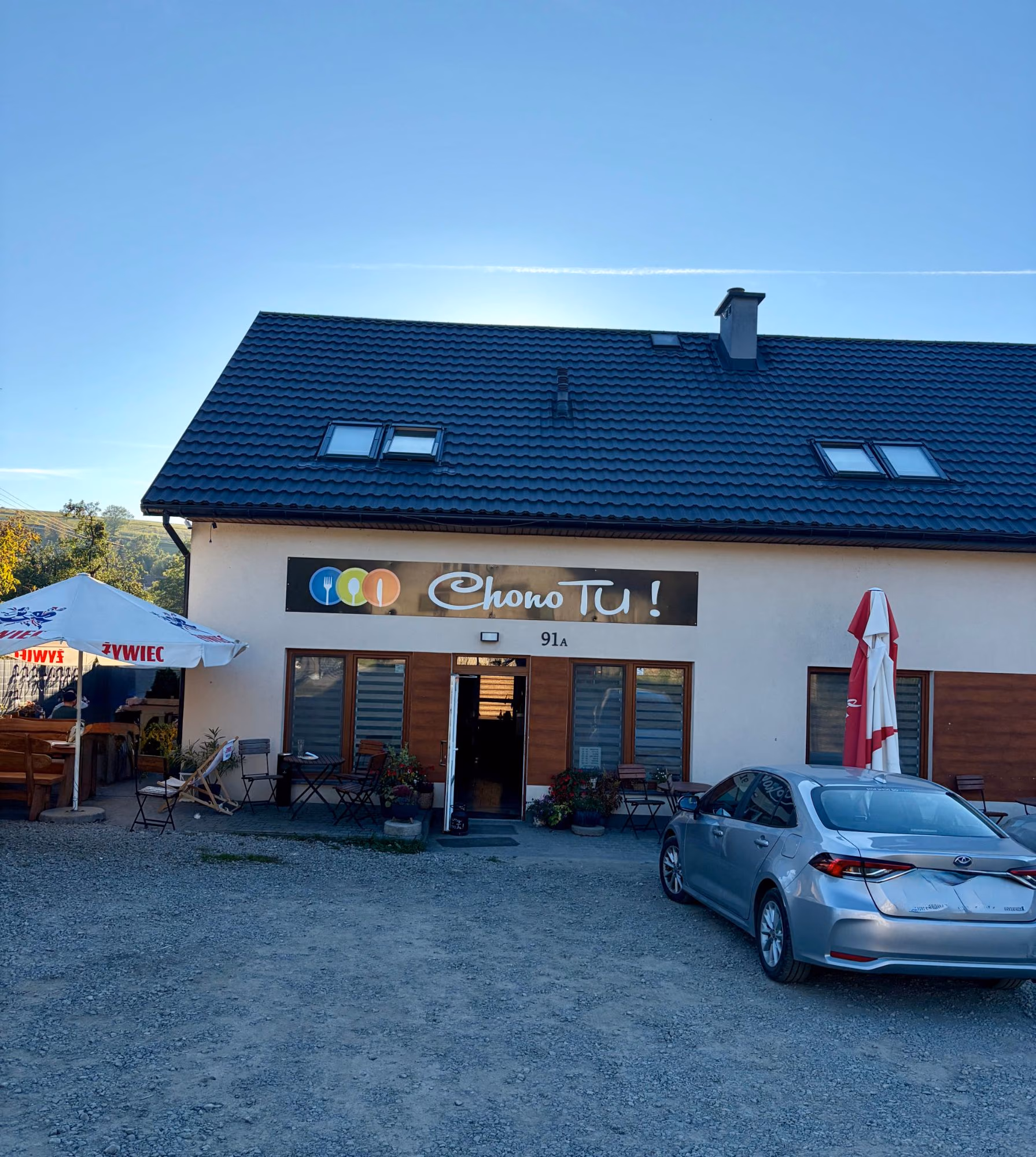 Jasny budynek pod ciemnym dachem z oknami dachowymi, nad wejściem szyld „Chono TU!”, przed nim stoliki i kwiaty, z lewej ogródek pod parasolem, z przodu żwirowy parking