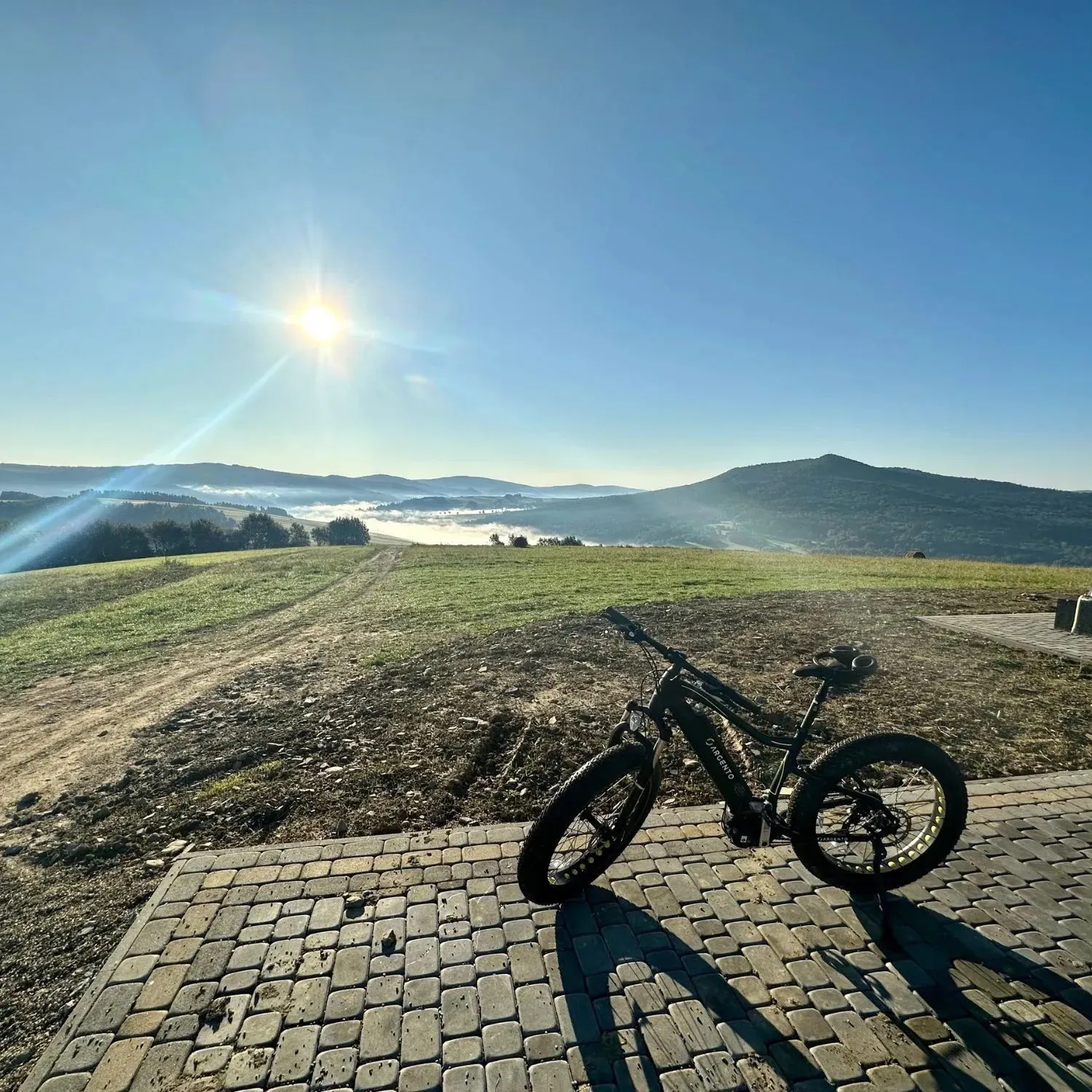Czarny elektryczny fatbike stoi na kostce na skraju łąki, w dole dolina zasnuta mgłą